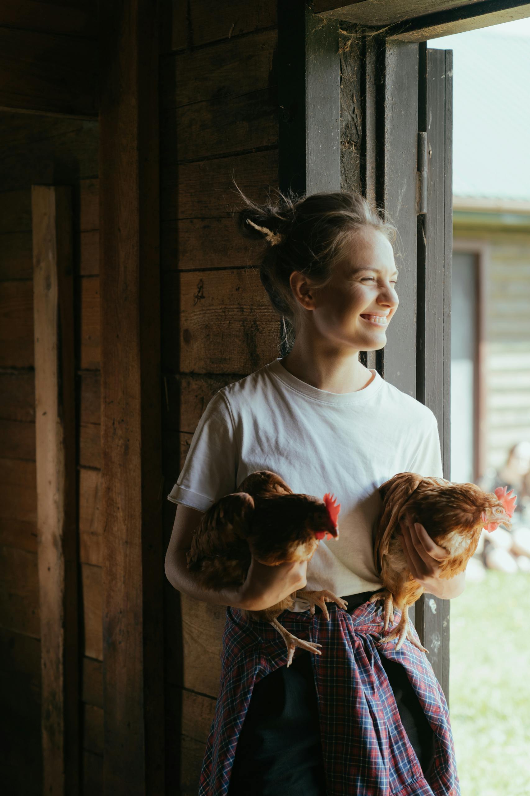 Happy woman holding chickens in a rustic barn, embracing farm life joyfully.